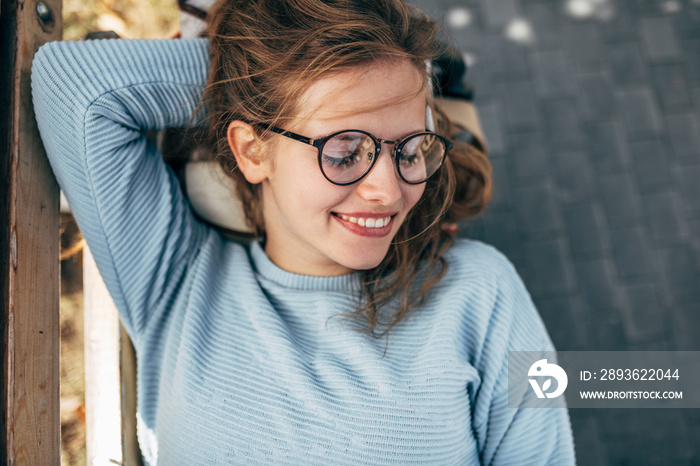 Gorgeous female in blue sweater, glasses, smiling with closed eyes, take a rest on the bench outside