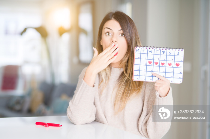 Young beautiful woman holding menstruation calendar at home cover mouth with hand shocked with shame