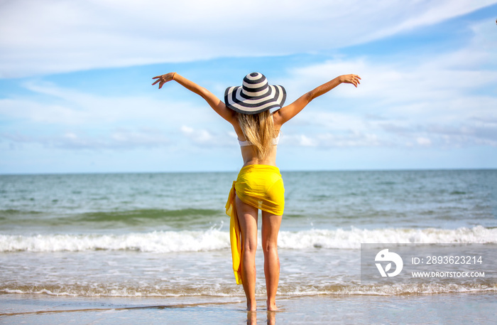 海滩度假。戴着太阳帽和比基尼的美女举起双臂站着的后视图