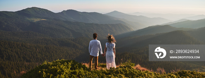 Back view of loving couple standing on grassy hill and holding hands while looking at beautiful moun