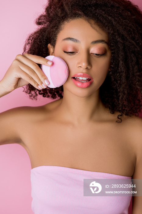 african american woman with closed eyes using silicone cleansing facial brush, isolated on pink