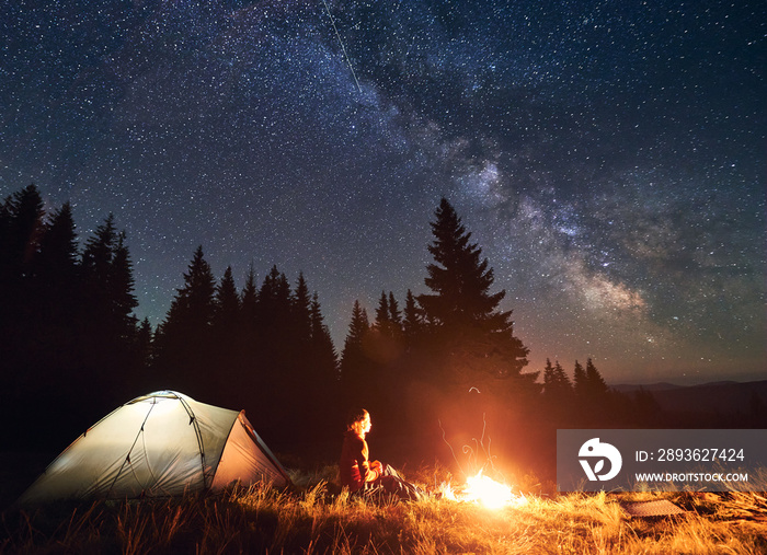 Rear view of woman hiker sitting by bright burning campfire near tent alone, thinking and enjoying b