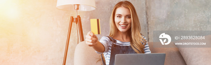 happy beautiful woman showing credit card and sitting on sofa with laptop
