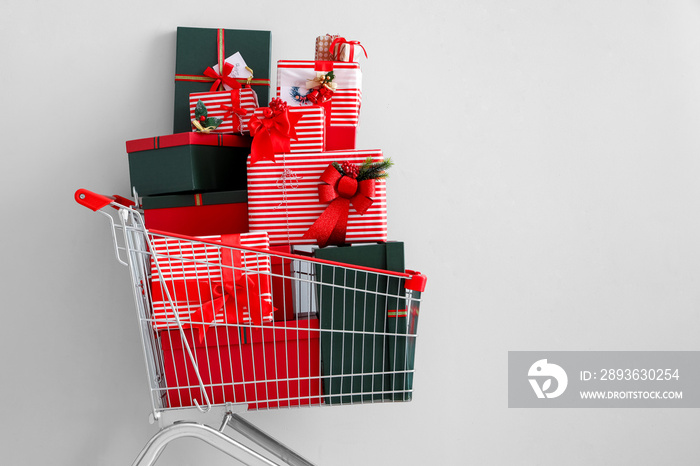 Shopping cart with beautiful Christmas gifts near light wall