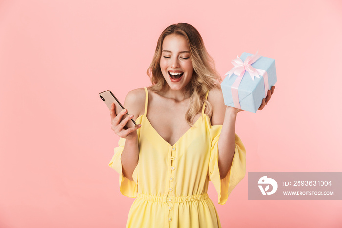 Excited emotional happy young blonde woman posing isolated over pink wall background holding present