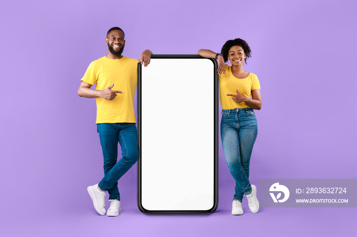 Cheery black couple pointing at smartphone on violet studio background, mockup for mobile app on whi