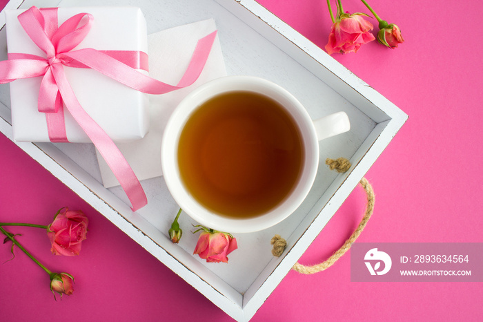 Tea in the cup and gift with pink bow on the white wooden tray on the dark pink  background.Top view