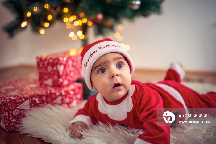 Cute baby in Christmas costume and gift box on floor at home