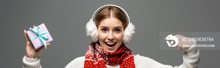 panoramic shot of excited girl in scarf and ear warmers pointing at christmas gift, isolated on grey