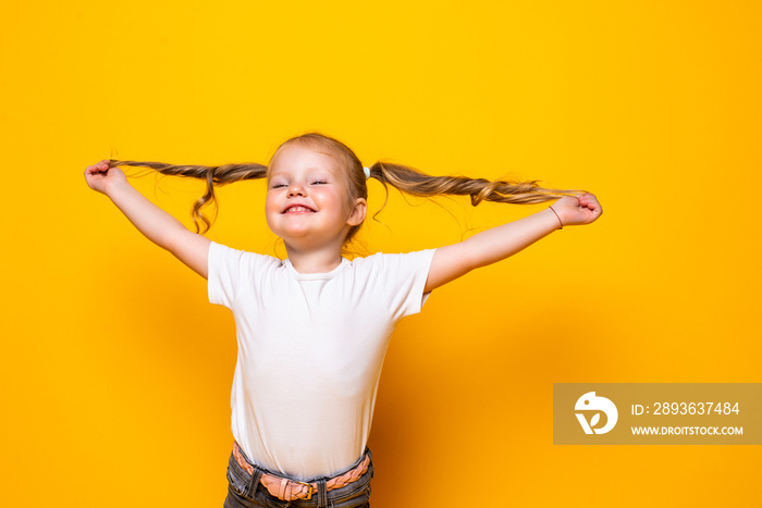 Joyful little girl playing with hair posing isolated on yellow background