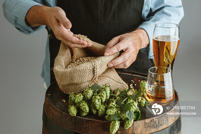 Close up of confident senior man brewer with self crafted beer in glass on wooden barrel on grey bac