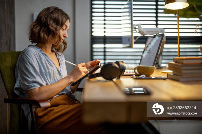 Young woman dressed casually having some creative work, drawing on a digital tablet, sitting at the 