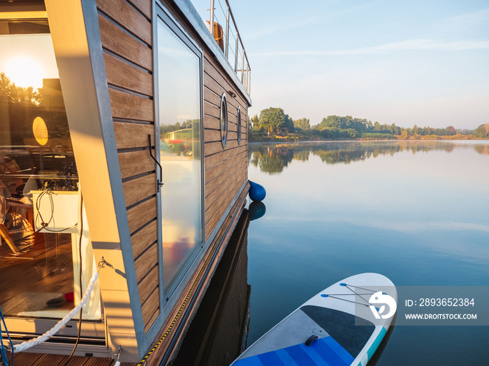 houseboat on a river in a early sunny morning moored on a pier with paddleboard. floating house is a