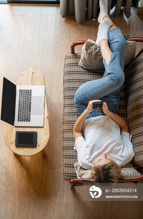女人躺在沙发上，桌上放着笔记本电脑和电子阅读器，一边使用智能手机。在线学习，
