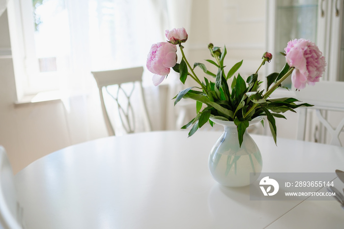 明亮的房间里，白色桌子上的圆形花瓶里摆放着粉红色的牡丹花和绿色的叶子
