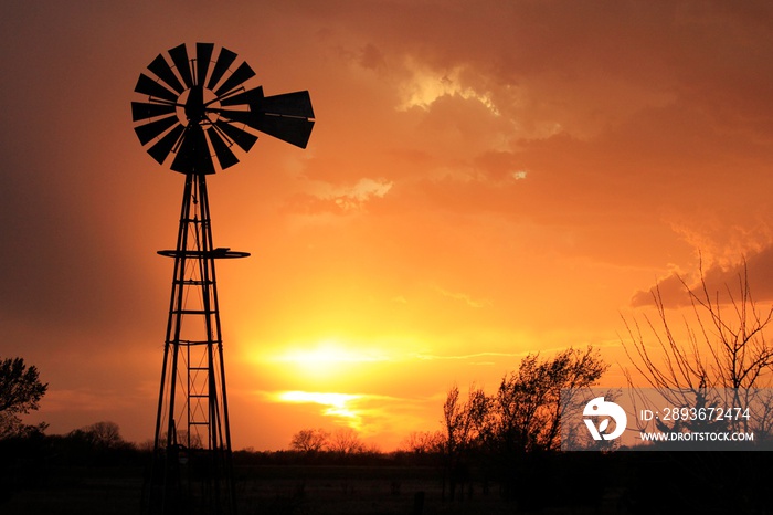 A Sunset with a Windmill Silhouette thats a blazing orange with trees out in the Country north of 