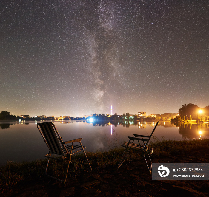 Two camping chairs on lake shore under beautiful night sky full of stars and Milky way, quiet water 