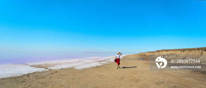 全景图美丽的风景，一个女人在盐粉色湖泊附近的海滩上行走