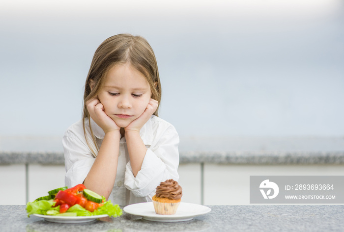 悲伤的小女孩在健康蔬菜和纸杯蛋糕之间做出选择。文本空间