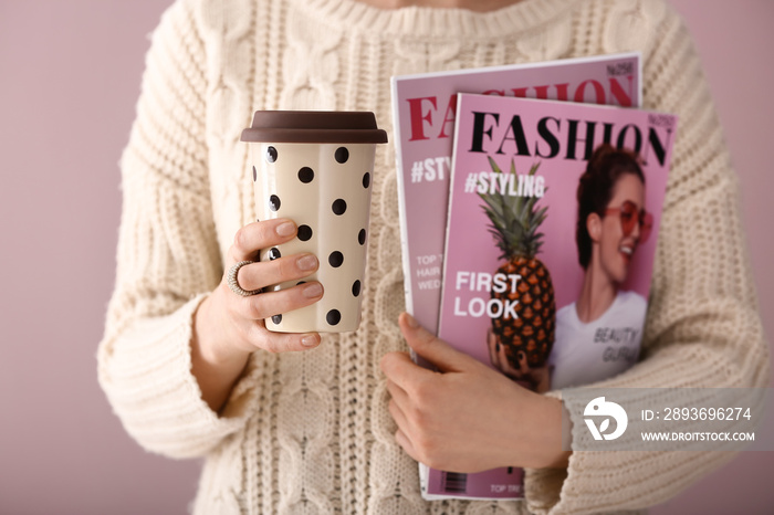 Young woman holding cup of coffee and magazines on color background, closeup