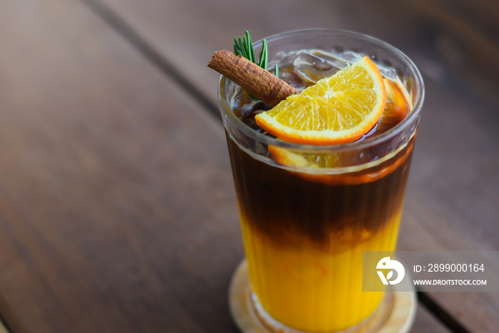 Top-view, Close-up of Iced Black coffee mixed with orange juice in glass topping with sliced oranges, rosemary leaves and cinnamon on top. on a wooden coaster placed on a wooden table.