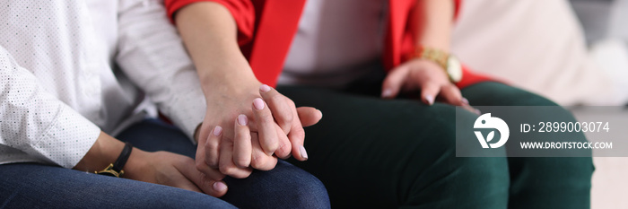 Two women tightly holding hands on couch at home close-up. Psychological support concept