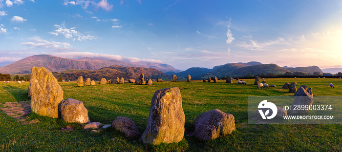 A panorama of Castlerigg stone circle looking south at sunset the dusk light painting the stones and casting their shadows across the grass, sheep graze in the circle wispy white clouds fleck the sky