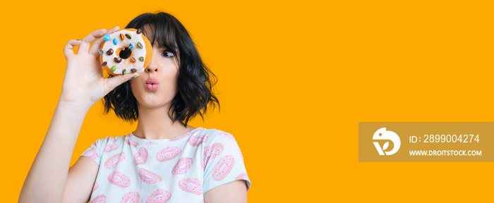 Charming brunette girl looking through a donut is feeling amazed posing on a yellow background with freespace