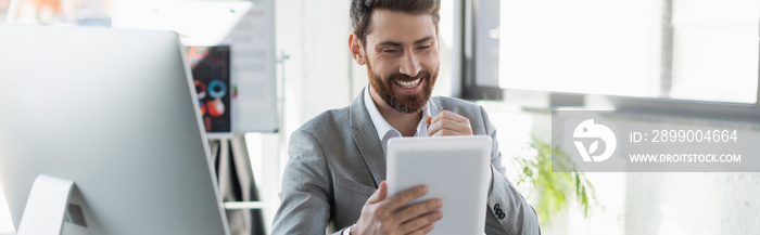 Smiling businessman holding digital tablet near computer monitor in office, banner.