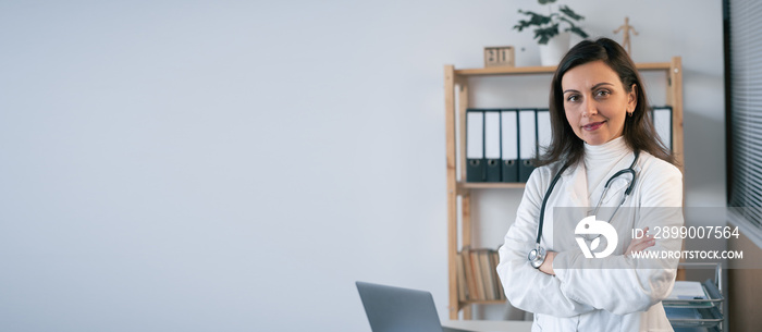 Female doctor with smile is looking at camera while standing with folded arms in medical clinic. Copy space.