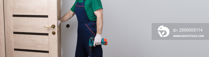 A male locksmith installs a door in an apartment. The guy works with a screwdriver. Panoramic banner.