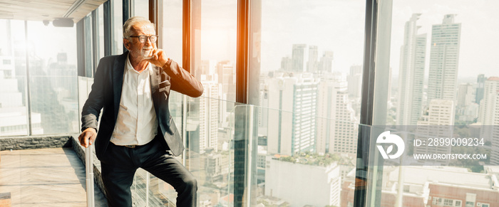 Retirement Man and insurance Concept.Portrait of Senior grey-haired businessman in suit standing and and looking through window the cityscape in office tower.