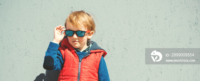 Stylish kid in trendy sunglasses. Kids fashion. Cute little blondy boy in red jacket standing over grey wall outdoors in sunny day. Autumn, spring kids fashion. Kids sunglasses. Hipster style.