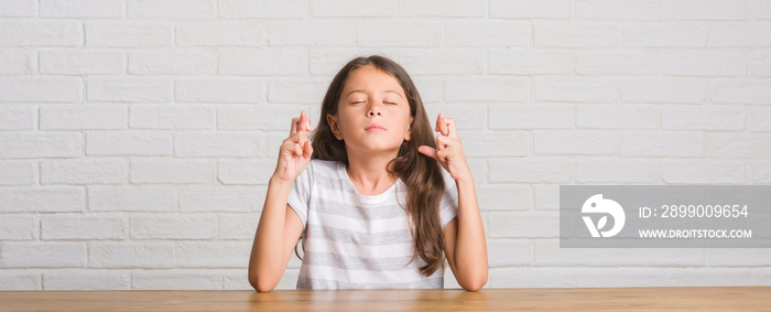 Young hispanic kid sitting on the table at home smiling crossing fingers with hope and eyes closed. Luck and superstitious concept.