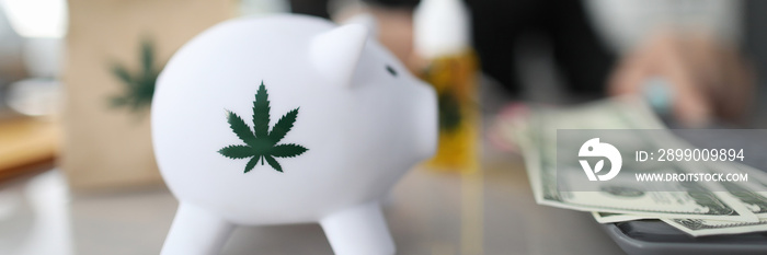 Close-up of piggy bank with green sign of cannabis. Paper package and bottle of cannabinoid oil with marijuana symbol on table. Cash on desk. Painkillers and medical marihuana concept