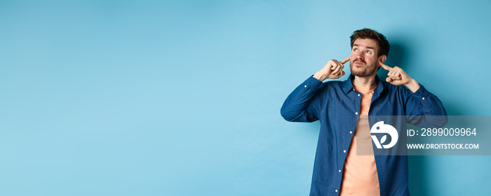 Annoyed young man block sound, shut ears with fingers and grimacing, disturbed by loud neighbours, looking up irritated, standing on blue background