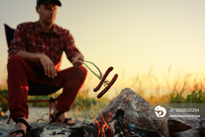 Young man roasting tasty sausage on a stick over the camp fire.