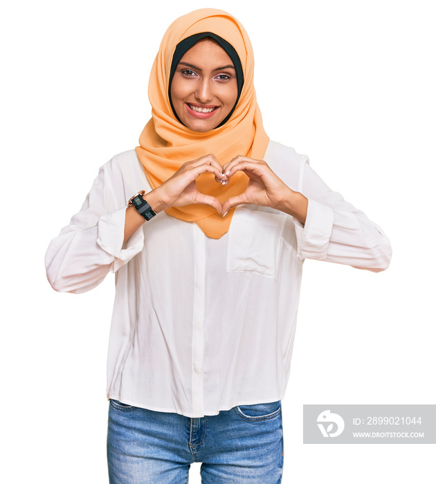 Young brunette woman wearing traditional islamic hijab scarf smiling in love showing heart symbol an