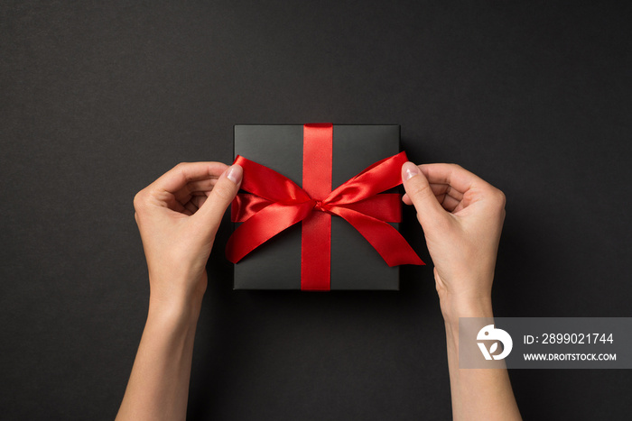 First person top view photo of hands tying red ribbon bow on black giftbox on isolated black backgro