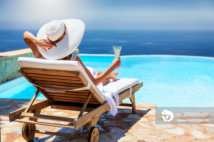A elegant woman in white dress and hat relaxes with a glass of champagne by the poolside on a sunny 