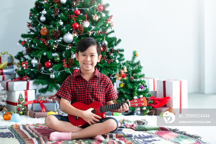 Asian kid sat in front of the Christmas tree with red ukulele, its Christmas time