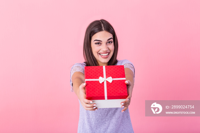 Happy brunette woman in casual holding gift box and looking at the camera while enjoys over pink bac