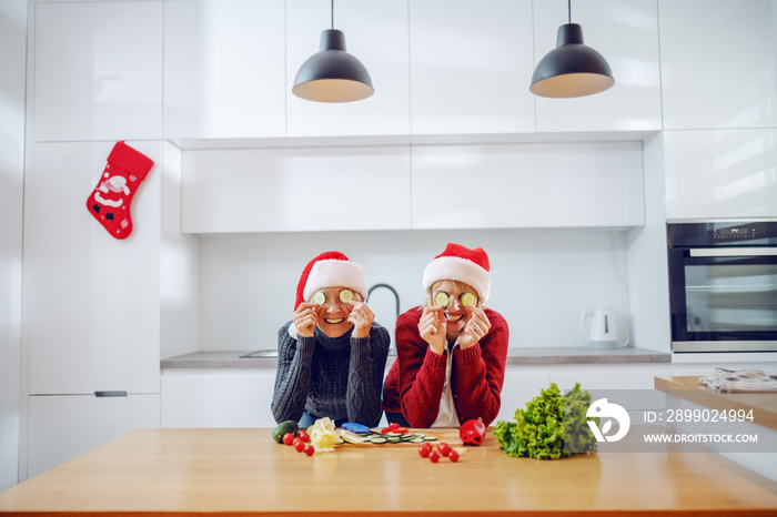 Playful caucasian woman and her mother holding cucumber slices in front of their eyes while leaning 