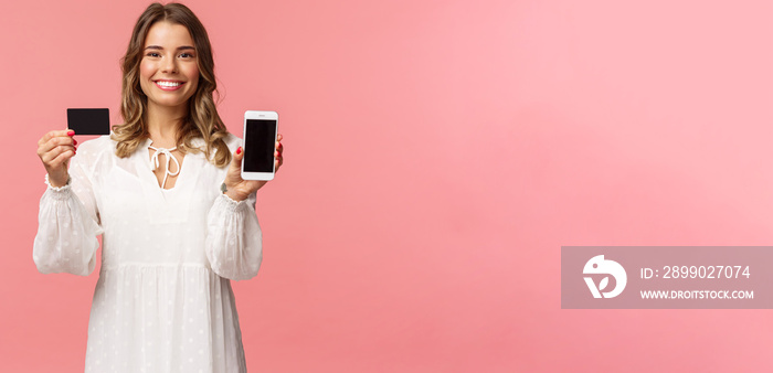 Portrait of satisfied cute blond girl in white dress, showing you her credit card and mobile phone a