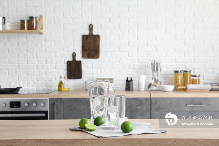 Jug and glasses with fresh water and lime on table in kitchen