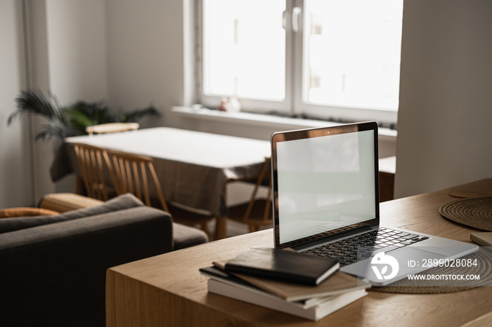 Laptop computer with blank screen, notebooks on wooden table in warm sunlight room. Aesthetic minima