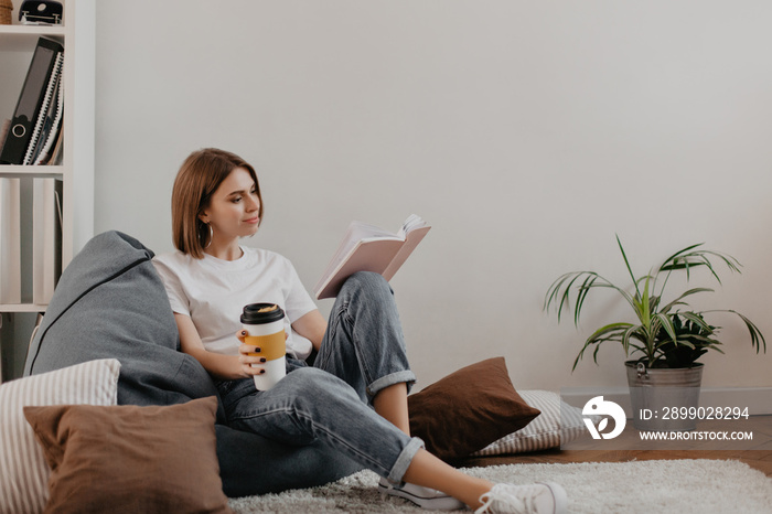 Full-length shot of young woman in stylish outfit. Girl enjoys book and coffee while sitting in cozy