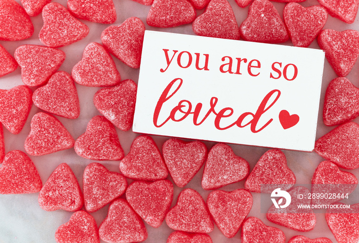 Overhead image of a you are loved sign surrounded by heart shaped red candies on a marble table. Ima