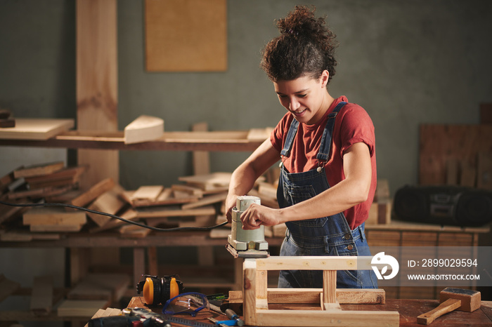 Young pretty female carpenter carefully smoothing wooden stool with electrical belt sander and smili