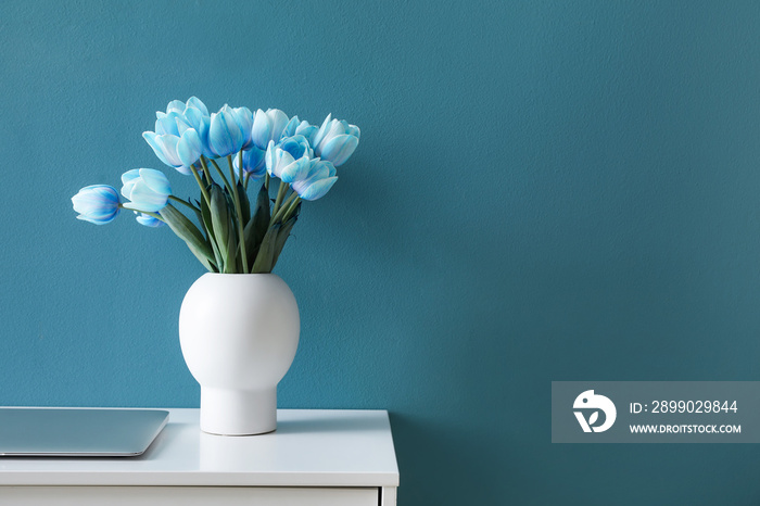 Vase with blue tulips and laptop on table near color wall